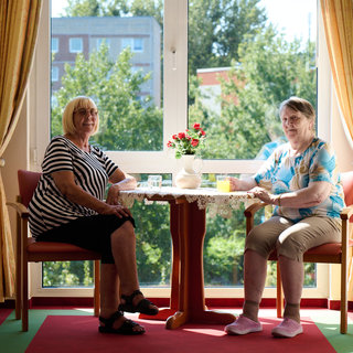 Zwei Frauen sitzen an einem Tisch in einem hellen Raum mit Blick auf einen Garten. Sie haben Getränke vor sich und wirken entspannt. Der Tisch ist mit einer Tischdecke und einer Vase mit Blumen dekoriert. Tageslicht strömt durch das Fenster.