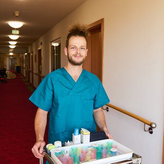 Ein junger Mann in blauer Arbeitskleidung steht lächelnd in einem langen Flur. Er schiebt einen Rollwagen, der mit verschiedenen Pflegeprodukten und -utensilien beladen ist. Die Wände des Flurs sind hell, und der Boden ist mit rotem Teppich ausgelegt.