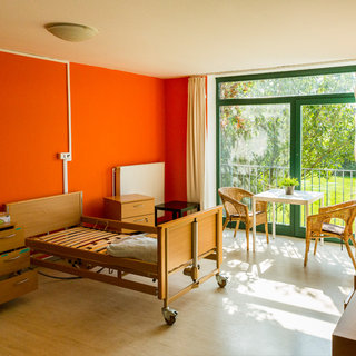 Ein helles Zimmer mit orangefarbener Wand. Im Vordergrund steht ein verstellbares Bett, daneben ein rollbarer Schrank. In der Ecke befindet sich ein Tisch mit zwei Stühlen, und große Fenster bieten Ausblick auf einen grünen Außenbereich mit Bäumen. Tageslicht strömt herein.