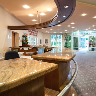 Ein Empfangsbereich mit einem glänzenden, runden Empfangstresen aus Marmor. Im Hintergrund sind Pflanzen und große Fenster zu sehen, die den Raum hell erleuchten. An der Wand hängen Bilder und eine Uhr ist sichtbar. Der Raum wirkt freundlich und einladend.