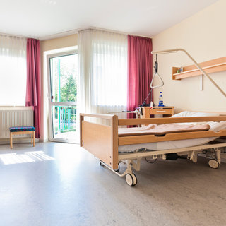 Ein helles Krankenzimmer mit einem Pflegebett, das auf Rädern steht. Neben dem Bett befindet sich ein Nachttisch mit einem Regal darüber. Am Fenster hängen leichte Vorhänge, und es gibt einen kleinen Tisch mit einem Stuhl. Tageslicht strömt durch die Fenster.