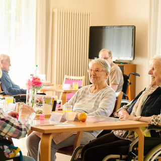 Eine Gruppe von älteren Menschen sitzt in einem hellen Raum. Einige sind in Rollstühlen, während andere an Tischen mit Getränken sitzen. Sie unterhalten sich und wirken freundlich und entspannt. Im Hintergrund ist ein Fernseher zu sehen.