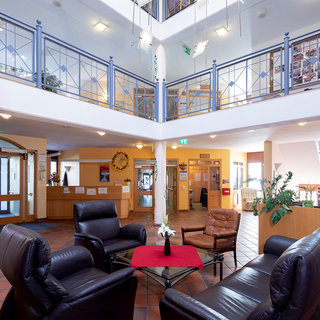 Eine helle Lobby mit einem offenen Atrium. Im Erdgeschoss stehen schwarze Ledersofas um einen kleinen Tisch mit einer Blume. Umgibt von Pflanzen und einem Empfangstresen. Helle Wände und große Fenster sorgen für eine freundliche Atmosphäre.