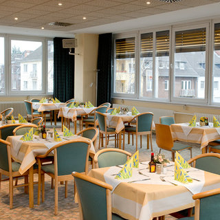 Das Bild zeigt ein Restaurant mit mehreren Tischgedecken. Jede Tischplatte ist mit einer weißen Tischdecke und Papierservietten in grünen und gelben Farben dekoriert. Es gibt Stühle um die Tische und große Fenster, die einen Blick ins Freie ermöglichen.