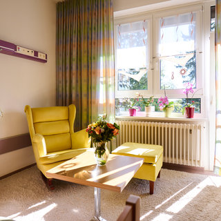 Ein helles Wohnzimmer mit einem gelben Sessel und einem kleinen Tisch. Auf dem Tisch steht eine Vase mit frischen Blumen. Große Fenster lassen viel Licht herein, während bunte Vorhänge die Umgebung auflockern. Im Hintergrund sind Pflanzen auf der Fensterbank zu sehen.