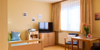 Ein helles, freundliches Zimmer mit einem Bett an der Wand, einem Fernseher auf einem Schrank und einem Tisch mit Stühlen. Vor dem Fenster hängen Vorhänge, und eine kleine Blumenvase mit Blumen steht auf dem Tisch. An der Wand sind Bilder rahmen.