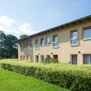 Ein mehrstöckiges Gebäude mit hellen Fassadenfenstern und einem Garten davor. Der Garten besteht aus einer schmalen Hecke und gepflegtem Gras. Im Hintergrund sind einige Bäume und ein blauer Himmel mit wenigen Wolken sichtbar.