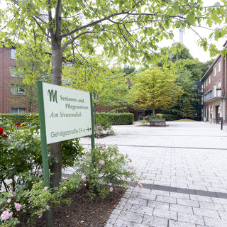 Ein Eingang zu einem Senioren- und Pflegezentrum, umgeben von einem gepflegten Garten mit Blumen und Bäumen. Ein großes Schild zeigt den Namen der Einrichtung und die Adresse an. Der Platz ist ruhig und lädt zum Verweilen ein.