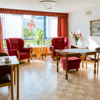 Ein heller Wohnraum mit großen Fenstern und roten Vorhängen. Zwei rote Sessel stehen gegenüber, ein Holztisch und Stühle sind im Raum verteilt. Auf einem Tisch stehen Kaffeetassen, und eine Vase mit Blumen schmückt die Ecke. Ein angenehmes, einladendes Ambiente.