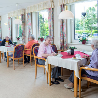 In einem hellen Raum sitzen ältere Frauen an Tischen und genießen ihre Getränke. Es gibt Blumen auf den Tischen und große Fenster, die natürliches Licht hereinlassen. Die Atmosphäre wirkt freundlich und einladend.