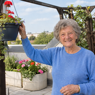Eine ältere Frau mit grauen Locken steht auf einer Terrasse und lächelt, während sie einen Blumentopf mit roten Geranien in der Hand hält. Im Hintergrund sind weitere blühende Pflanzen in Töpfen zu sehen, umgeben von Holzgittern. Der Himmel ist blau mit einigen Wolken.