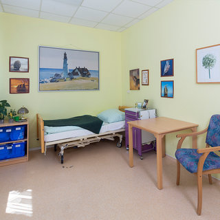 Ein helles Krankenzimmer mit einem Bett, einer Kommode und einem Tisch. An der Wand hängen Bilder, darunter ein Landschaftsbild mit einem Leuchtturm. Links steht ein kleiner Tisch mit einer Pflanze. Der Boden ist neutral und es gibt Tageslicht.