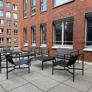 Eine Terrasse mit mehreren schwarzen Stühlen und Tischen aus Metall. Die Wände im Hintergrund sind aus roten Ziegeln, und große Fenster lassen Tageslicht hinein. Die Gestaltung wirkt modern und einladend.