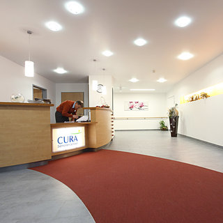 Die Empfangshalle eines Gesundheitszentrums mit einem modernen Empfangstresen. Ein Mitarbeiter steht am Tresen und ist mit administrativen Aufgaben beschäftigt. Der Fußboden ist grau und ein roter Läufer führt durch den Raum. An der Wand hängt ein Kunstwerk, und Pflanzen stehen dekorativ im Raum.