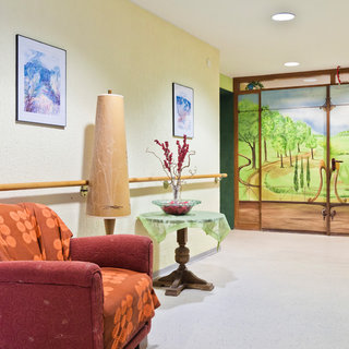 Ein blühender Flur mit einer bequemen roten Couch und einer Stehlampe. Auf einem Tisch stehen eine Vase mit Pflanzen und ein grünes Tuch. An der Wand befinden sich Bilder, und eine große bemalte Tür zeigt eine Landschaft mit Bäumen und Hügeln.