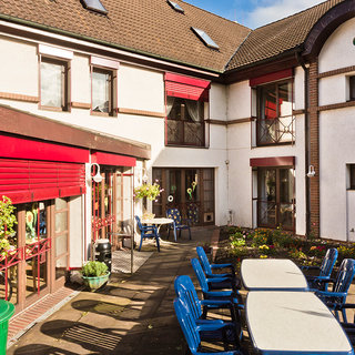 Eine sonnige Innenhofansicht eines Gebäudes mit roten Fensterläden. Im Vordergrund stehen mehrere blaue Stühle und ein Tisch. Um den Tisch herum sind Blumen und Pflanzen angeordnet. Das Gesamtbild vermittelt eine einladende, entspannende Atmosphäre.