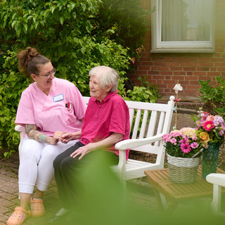 Eine Pflegekraft sitzt auf einer Bank neben einer älteren Dame im Freien. Beide lächeln und unterhalten sich freundlich. Neben ihnen steht ein Korb mit bunten Blumen. Die Umgebung ist grün und einladend.