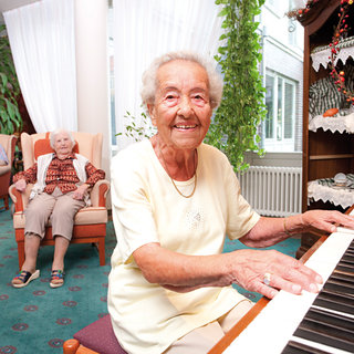 Eine ältere Frau sitzt an einem Klavier und lächelt, während sie spielt. Im Hintergrund sind zwei weitere ältere Frauen auf bequemen Sesseln zu sehen. Der Raum ist hell und mit Pflanzen dekoriert, was eine freundliche Atmosphäre schafft.