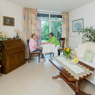 Zwei Personen sitzen am Tisch in einem hellen Wohnzimmer. Die eine Person trägt ein rosa T-Shirt, die andere ein grünes. Auf dem Tisch stehen eine Blumenvasen und Obst. Im Hintergrund ist ein Fenster mit Ausblick auf das Grüne.