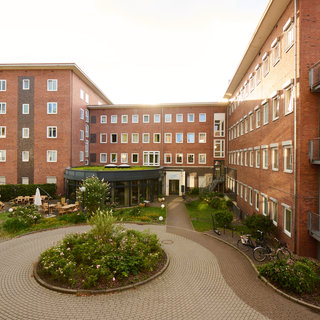 Das Bild zeigt einen Innenhof eines Mehrfamilienhauses mit mehreren Backsteingebäuden. Im Vordergrund sind gut angelegte Gartenflächen mit Pflanzen und Sitzmöglichkeiten zu sehen. Der Innenhof hat einen gepflasterten Weg, der sich sanft in die Landschaft einfügt.