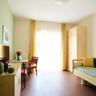 Das Bild zeigt ein helles, modernes Zimmer mit einem Einzelbett, einem Schreibtisch und einem Stuhl. Große Fenster mit Vorhängen lassen Tageslicht herein. An der Wand sind ein Spiegel und ein Bild zu sehen. Pflanzen und farbige Kissen ergänzen die gemütliche Atmosphäre.