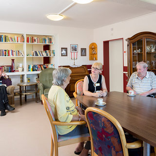 In einem Raum sitzen vier Erwachsene um einen Tisch. Zwei Frauen und zwei Männer führen ein Gespräch, während einer der Männer im Hintergrund in einem Sessel sitzt und liest. Ein Bücherregal und ein Vitrinenschrank sind im Raum sichtbar.