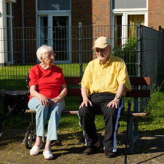 Zwei Senioren sitzen auf einer Bank in einem Garten. Die Frau trägt ein rotes Shirt und die Männer ein gelbes. Sie unterhalten sich freundlich, während im Hintergrund ein Rollator steht. Es ist ein sonniger Tag, und die Atmosphäre wirkt entspannt und einladend.