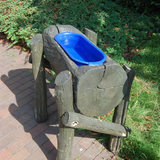 Ein Holzwassertrog mit blauer Schüssel steht auf einem gepflasterten Weg. Die Ränder sind grob bearbeitet, und die Beine sind stabil. Die Umgebung besteht aus Gras und Sträuchern.