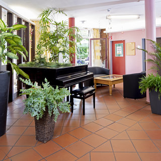 Der Raum zeigt ein einladendes Foyer mit einem Klavier und mehreren Pflanzen in dekorativen Töpfen. Rechts stehen zwei schwarze Sessel und ein Holztisch. Die Wände sind in sanften Farben gestrichen, und große Fenster sorgen für viel natürliches Licht.