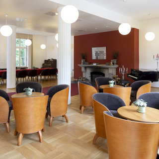 Ein hell erleuchteter Raum mit mehreren runden Tischen und eleganten Stühlen. Im Hintergrund steht ein Klavier, und es gibt einen Kamin mit Kerzen. Große Fenster lassen viel Tageslicht herein, und dekorative Pflanzen sind in den Ecken platziert.