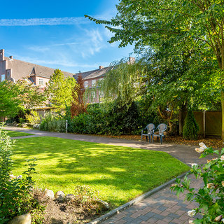 Ein gepflegter Garten mit grüner Rasenfläche, umgeben von Bäumen und Sträuchern. In der Mitte befinden sich zwei Stühle auf einem gepflasterten Weg. Im Hintergrund sind Wohngebäude sichtbar unter einem blauen Himmel mit leichten Wolken.