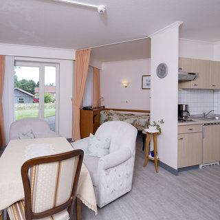 Ein gemütlicher Raum mit einer kleinen Küche und einer Essecke. Der Tisch ist mit einer Tischdecke gedeckt, während zwei Sessel und ein Klavier den Raum gemütlich gestalten. Große Fenster lassen viel Licht herein und bieten einen Blick ins Freie.