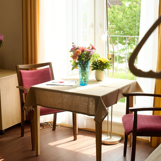 Ein heller Raum mit einem Tisch, der mit einer Tischdecke bedeckt ist. Auf dem Tisch stehen ein blauer Blumenvasen mit bunten Blumen und ein paar Unterlagen. Zwei Stühle sind um den Tisch herum angeordnet. Eine Fensterfront ermöglicht den Blick ins Freie.