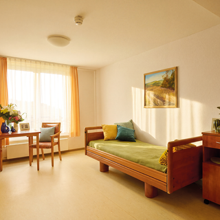 Ein helles, freundliches Zimmer mit einem Einzelbett, einem Tisch mit Stühlen und einem Nachttisch. An einem Fenster hängen orangene Vorhänge, und auf dem Tisch stehen Blumen. An der Wand hängt ein Landschaftsgemälde.