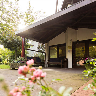 Eine Eingangsansicht eines Gebäudes mit einem überdachten Bereich und großen Fenstern. Der Boden ist gepflastert, und im Vordergrund wachsen rosafarbene Blumen. Das Licht der Sonne fällt durch das Dach und beleuchtet den Eingangsbereich.