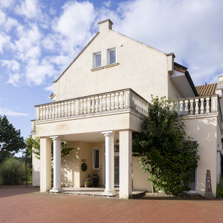 Eine helle, zweigeschossige Villa mit einem Balkon und Säulen am Eingang. Grüne Pflanzen wuchern um das Haus, und im Vordergrund ist ein gepflasterter Bereich sichtbar. Der Himmel ist teilweise bewölkt, was eine freundliche Atmosphäre vermittelt.