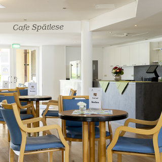 Das Bild zeigt das Innere eines Cafés mit dem Namen „Café Spätele“. Mehrere Holzstühle mit blauen Polstern umgeben runde Tische. Im Hintergrund sind eine helle Theke und eine Blumendekoration zu sehen. Das Ambiente wirkt freundlich und einladend.