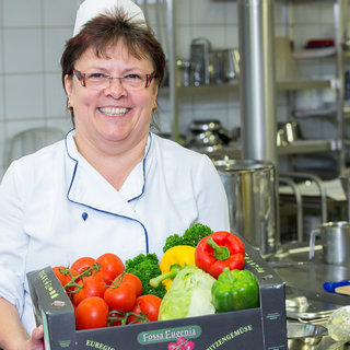 Eine lächelnde Köchin trägt einen Kasten mit frischem Gemüse, darunter Tomaten, Brokkoli, Paprika und Zucchini. Im Hintergrund sind Küchengeräte und Arbeitsflächen zu sehen, die eine geschäftige Küchenatmosphäre vermitteln.