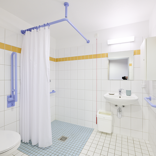Ein barrierefreies Badezimmer mit weißen Fliesen und einem blauen Fliesenboden. Eine Duschkabine mit einem weißen Vorhang, Haltegriffen in Blau und einem Waschbecken mit Spiegel sind vorhanden. Ein WC und ein kleiner Mülleimer sind ebenfalls zu sehen.