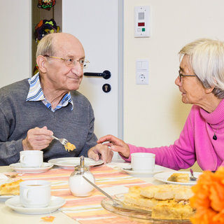 Ein älteres Paar sitzt an einem Tisch und genießt gemeinsam Gebäck und Kaffee. Der Mann, der einen grauen Pullover trägt, isst mit einem Löffel, während die Frau in einem pinken Pullover ihm anlächelt und ihn anfasst. Eine freundliche, gesellige Atmosphäre ist sichtbar.