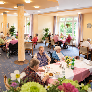 Ein Speisesaal mit vielen älteren Menschen, die an Tischen sitzen und essen. Einige nutzen Rollatoren. Die Tische sind mit rosa Tischtüchern gedeckt und es gibt Pflanzen sowie Getränke auf den Tischen. Die Atmosphäre ist freundlich und einladend.