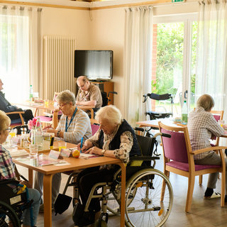 Eine Gruppe älterer Menschen sitzt in einem hellen Raum mit großen Fenstern. Einige Personen arbeiten an Tischen, während andere in Rollstühlen sitzen. Es herrscht eine freundliche Atmosphäre, und das Licht fallt auf die verschiedenen Aktivitäten.