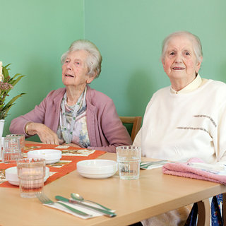 Zwei ältere Frauen sitzen an einem Tisch, der mit Geschirr und Gläsern gedeckt ist. Eine Frau trägt einen lila Pullover und schaut zur Seite, während die andere Frau in einem weißen Pullover lächelt. Im Hintergrund steht eine kleine Pflanze in einer Vase.