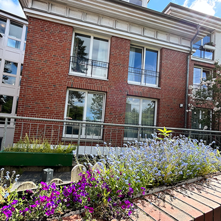 Blühende Blumenbeete mit lila und blauen Blumen sind im Vordergrund zu sehen, vor einem modernen, dreigeschossigen Gebäude aus rotem Backstein. Das Gebäude hat große Fenster und schlichte Balkone. Im Hintergrund sind weitere Bäume und Blumen sichtbar, bei sonnigem Wetter.