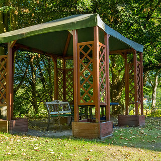 Eine holzrahmige Pergola mit grünem Dach steht in einem Garten. Unter dem Dach befindet sich eine Bank und ein Tisch. Umgeben von Bäumen und auf Grasboden, vermittelt die Szene einen einladenden und ruhigen Rückzugsort in der Natur.
