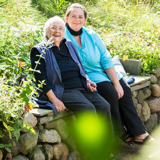 Zwei Frauen sitzen auf einer Steinbank in einem Garten. Die ältere Frau trägt eine Brille und lächelt. Die jüngere Frau hat lange Haare und lächelt ebenfalls. Um sie herum wachsen Pflanzen und die Umgebung scheint sonnig und angenehm zu sein.