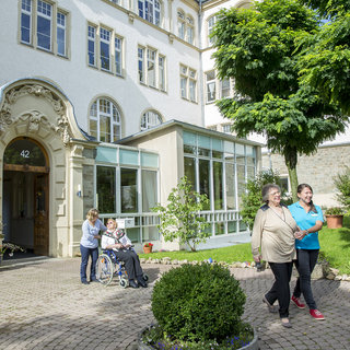 Zwei Pflegerinnen begleiten eine ältere Frau spazierend im Freien. Eine weitere ältere Dame sitzt im Rollstuhl neben einem Gebäude mit großen Fenstern. Der Garten ist gepflegt und mit Pflanzen dekoriert.