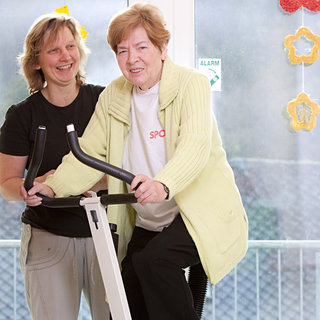 Eine ältere Frau trainiert an einem Fitnessgerät, während eine jüngere Trainerin sie unterstützt und ermutigt. Die Umgebung ist hell und freundlich, mit einer großen Fensterfront im Hintergrund.