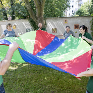 Eine Gruppe von fünf Personen steht im Freien und hält ein großes, buntes Tuch mit rot, grün, blau und gelb in den Händen. Sie stehen im Kreis und scheinen ein Spiel zu spielen, das gemeinschaftliche Aktivität und Freude ausdrückt. Im Hintergrund sind Bäume und eine Wand mit bunten Bildern sichtbar.