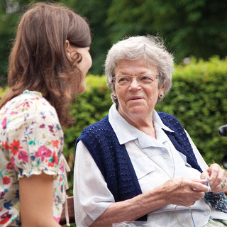 Zwei Frauen sitzen im Freien auf einer Bank. Die ältere Frau mit grauen Haaren und Brille scheint freundlich zuzuhören und zu lächeln, während die jüngere Frau mit langen, braunen Haaren sie anspricht. Im Hintergrund ist grünes Laub zu sehen.